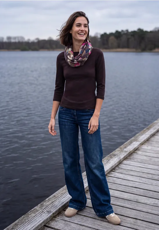 Eine lächelnde Frau mit Schal und Jeans steht auf einem Holzsteg am ruhigen See im Wald.
