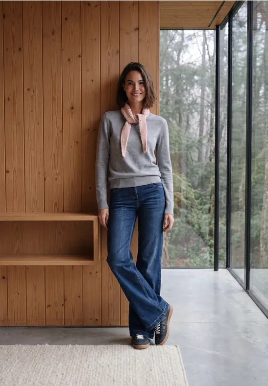 Eine Frau lehnt lässig in Jeans und grauem Pulli an einer Holz­wand mit großem Fenster und Waldsicht.