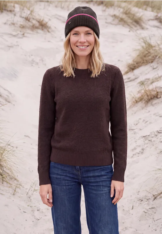 Eine Frau mit Strickmütze und Pullover lächelt am Dünenstrand in Jeans.