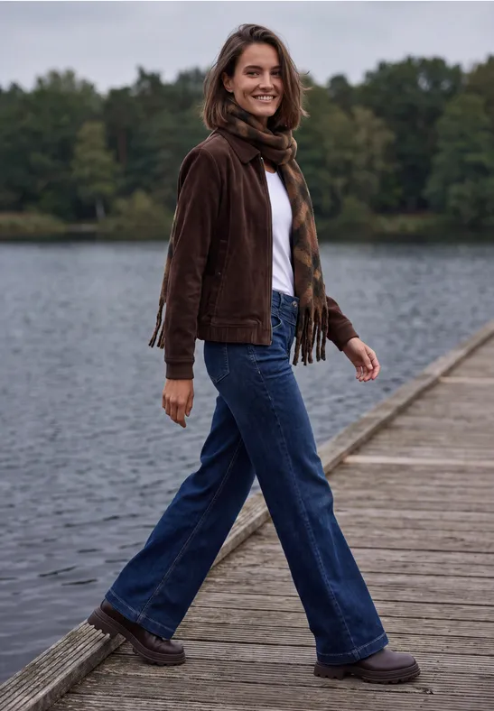 Une femme en veste brune et jean marche en souriant sur une jetée en bois au bord du lac.