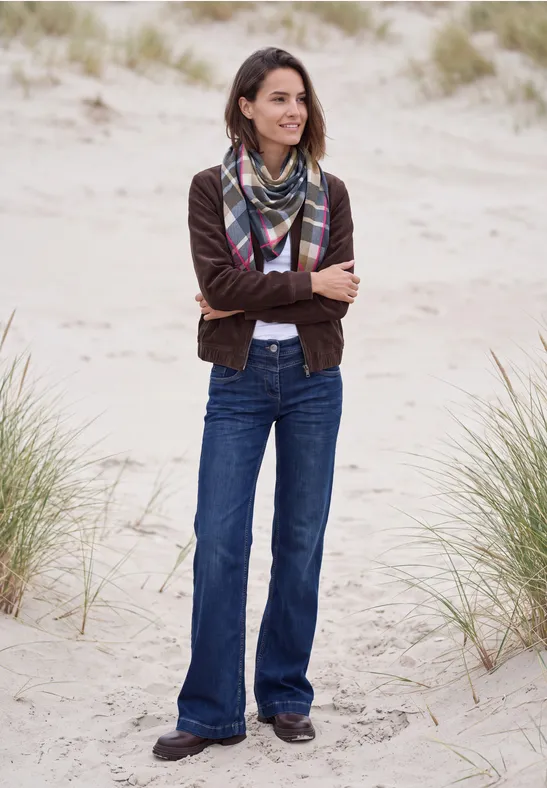Eine Frau steht in Jeans, Jacke und Schal mit verschränkten Armen lächelnd auf sandigen Dünen.