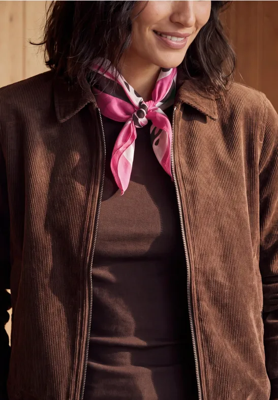 Personne en veste marron et top assorti, portant un foulard rose noué autour du cou.