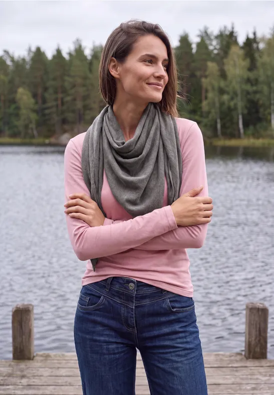 Een glimlachende vrouw in roze trui en sjaal staat aan de oever van een meer voor een bos.