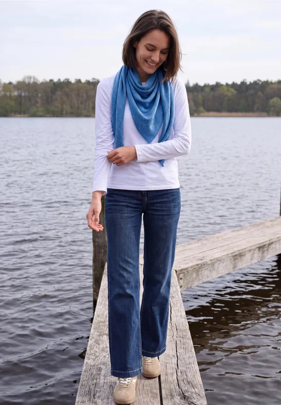 Une femme en jean, haut à manches longues et foulard bleu se tient souriante sur une jetée au bord d'un lac.
