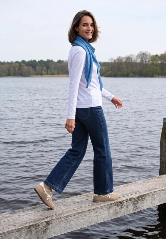 Femme avec écharpe bleue, jean et baskets marchant sur une jetée au bord du lac, souriant en arrière.