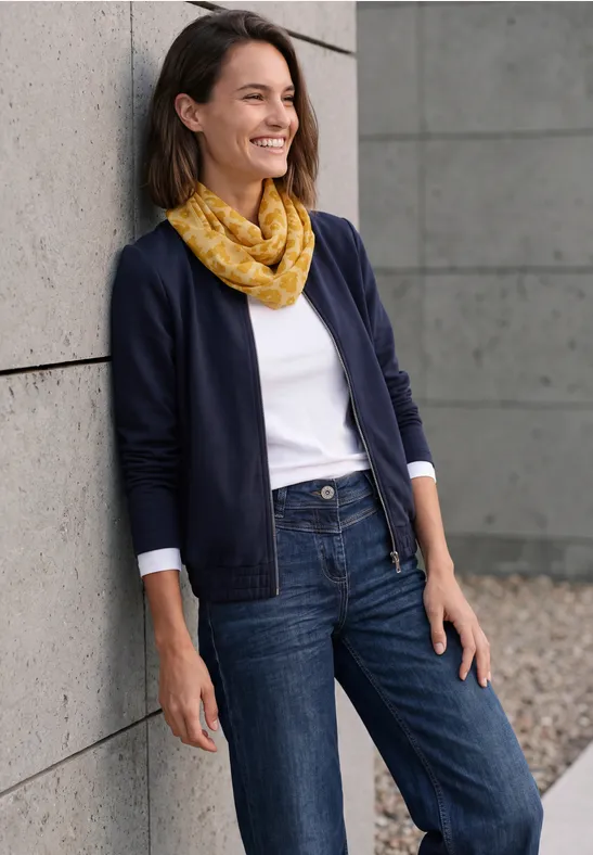 Eine lächelnde Frau lehnt im Freien an einer Wand, trägt Jeans, weißes Shirt, dunkelblauen Cardigan und gelben Schal.