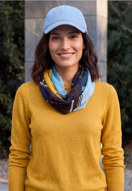 Eine lächelnde Frau mit gelbem Pullover, gemustertem Schal und hellblauer Mütze im Freien.