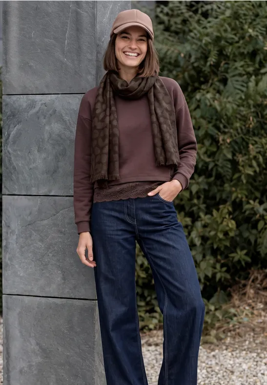Eine lächelnde Frau lehnt sich an eine Steinwand, trägt Mütze, Schal, braunen Pullover und blaue Jeans.