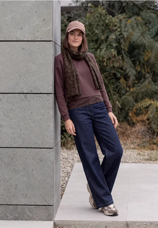 Eine junge Frau lehnt draußen an einer Wand, trägt Mütze, Schal, Pullover, blaue Hosen und Sneaker.
