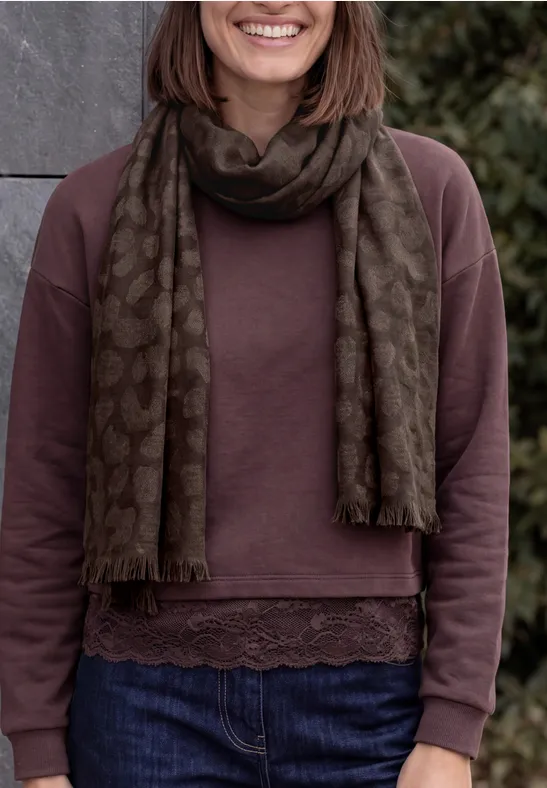Frau mit olivgrün gemustertem Schal, lila Pullover mit Spitzensaum und blauen Jeans.