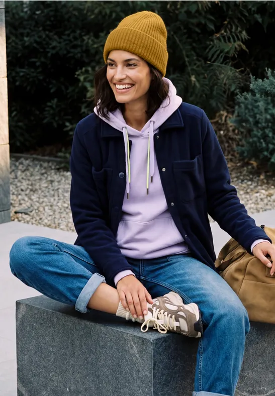 Junge Frau in Mütze, Hoodie, Jeans und Sneakers sitzt lächelnd im Freien auf einer Mauer.