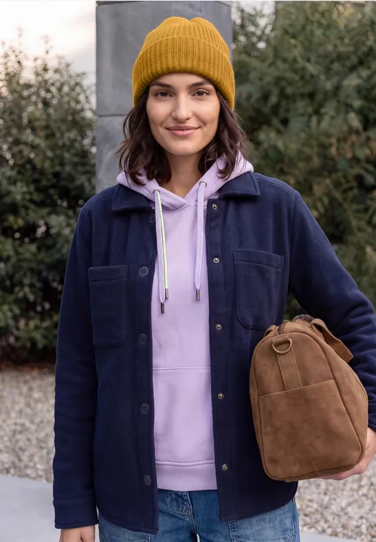 Junge Frau mit gelber Mütze und lila Kapuzenpullover trägt eine braune Tasche vor Grünpflanzen.