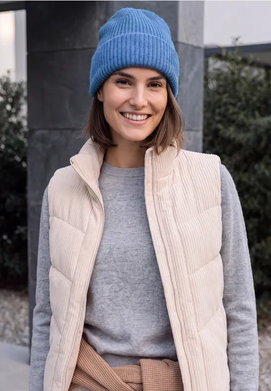 Een glimlachende vrouw met een blauwe muts, beige bodywarmer en grijze trui buiten.
