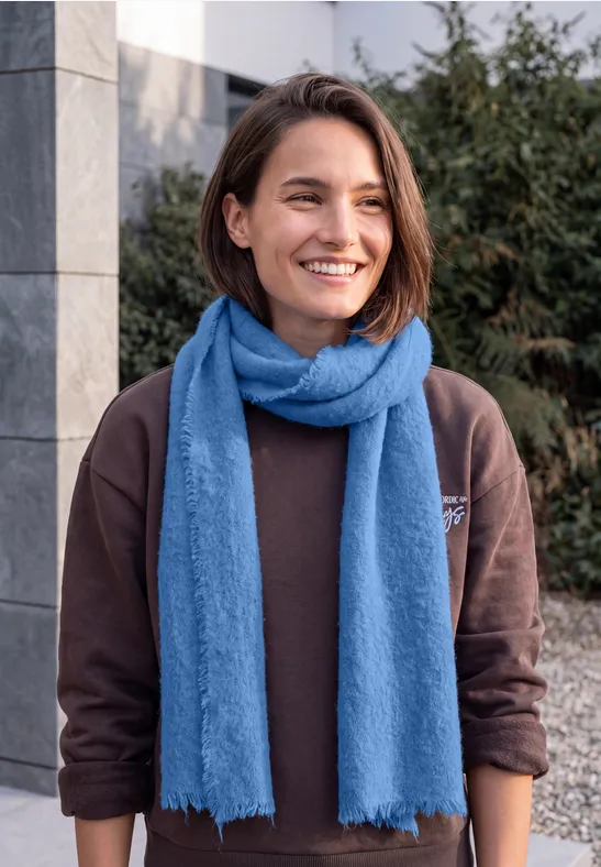 Junge Frau mit braunem Pullover und blauem Schal lächelt im Freien vor modernem Gebäude.
