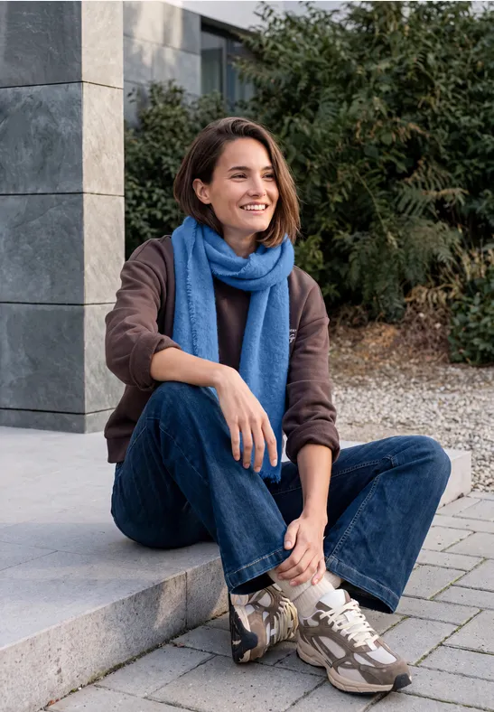Eine lächelnde Frau mit blauem Schal und Jeans sitzt entspannt auf einer Stufe vor einem modernen Gebäude.