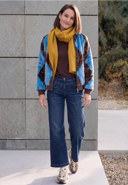 Eine Frau steht vor einer Betonwand, trägt bunte Patchworkjacke, gelben Schal, Jeans und Halbschuhe.