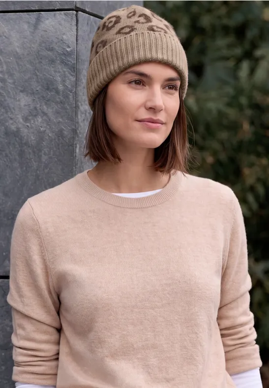 Junge Frau im beigen Pullover und gestrickter Mütze steht vor einer Steinwand, dahinter grüne Bäume.