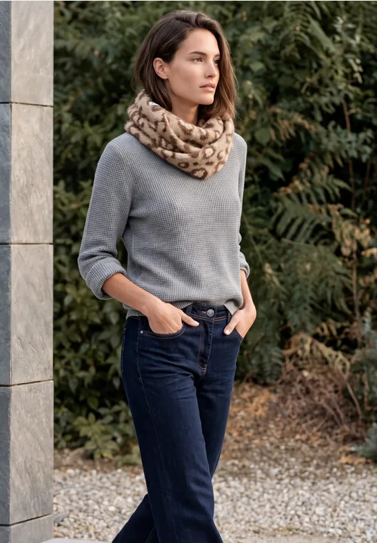 Frau in grauem Pullover und Schal, dunkle Jeans, Hände in Taschen, steht draußen an Säule.