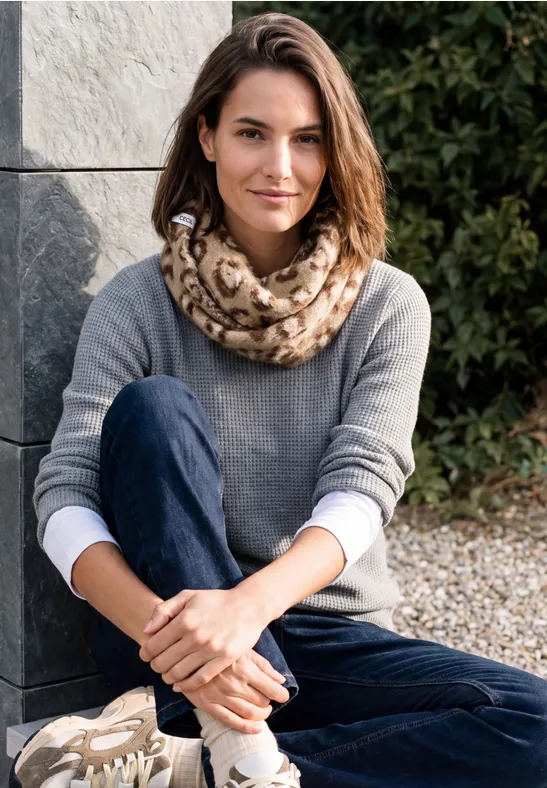 Eine Frau sitzt im Freien auf Kies, trägt grauen Pullover, Schal mit Leopardenmuster und Jeans.