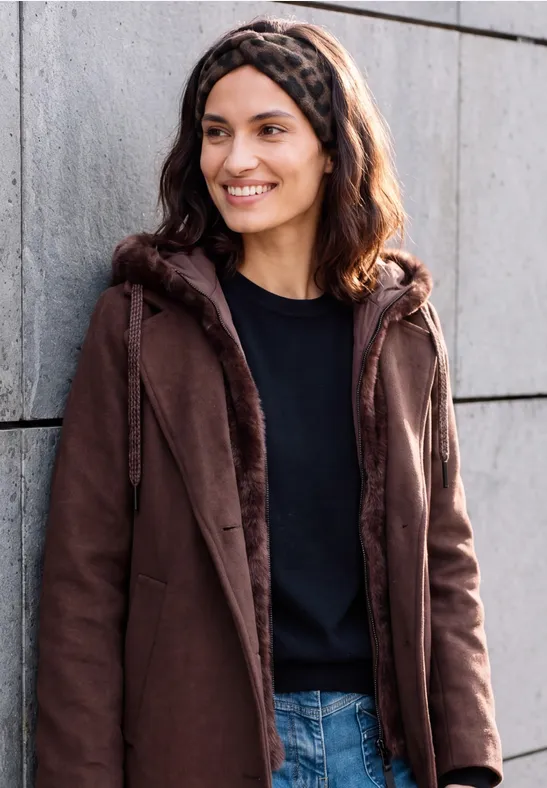Een glimlachende vrouw in een bruine jas, zwart shirt en jeans leunt tegen een grijze muur.