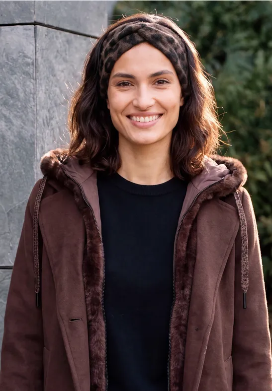Een lachende vrouw in een bruine jas, zwart shirt en haarband voor een stenen muur.