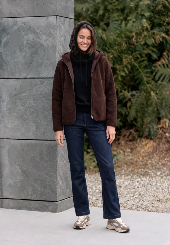 Vrouw in een bruine jas, jeans en sneakers leunt glimlachend tegen een grijze muur buiten, omringd door groen.