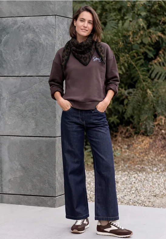 Frau in Pullover, Halstuch und breiten Jeans steht mit Händen in Taschen lässig vor Steinwand und Grün.