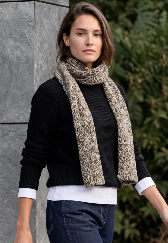 Frau in schwarzem Pullover, Jeans und beiger Strickschal posiert vor einer Natur- und Steinmauerkulisse.