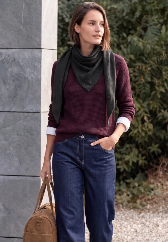 Frau in Jeans, weinrotem Pullover und schwarzem Schal trägt Tasche und lehnt an Wand.