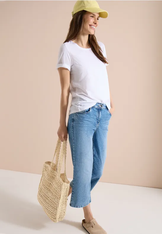 Frau in weißem T-Shirt, hellblauen Jeans, Strohtasche und Cap vor beigem Hintergrund