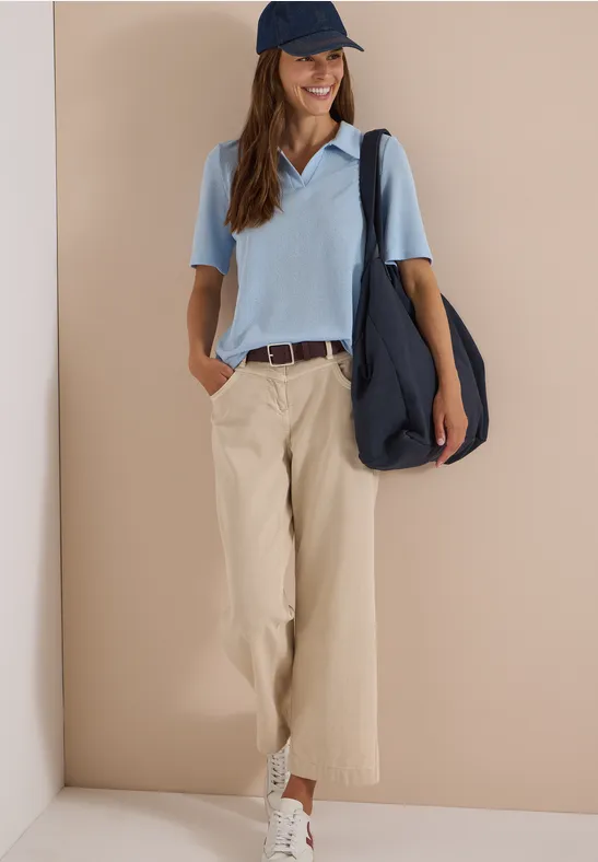 Frau trägt hellblaues Poloshirt, beige Stoffhose, weiße Sneaker, blaue Cap und große Umhängetasche.