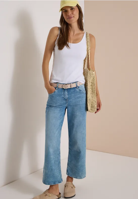 Eine Frau trägt ein weißes Tanktop, weite Jeans, beigen Gürtel, beige Schuhe, Strohtasche und gelbe Cap.