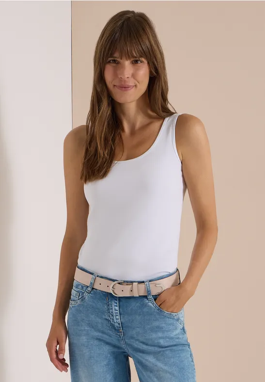 Junge Frau im weißen Tanktop und Jeans mit rosa-grauem Gürtel, lächelnd vor neutralem Hintergrund.