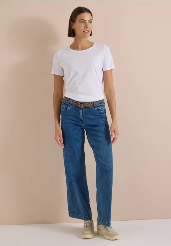 Frau in weißem T-Shirt, weiten Jeans und Sneakers vor beigem Hintergrund