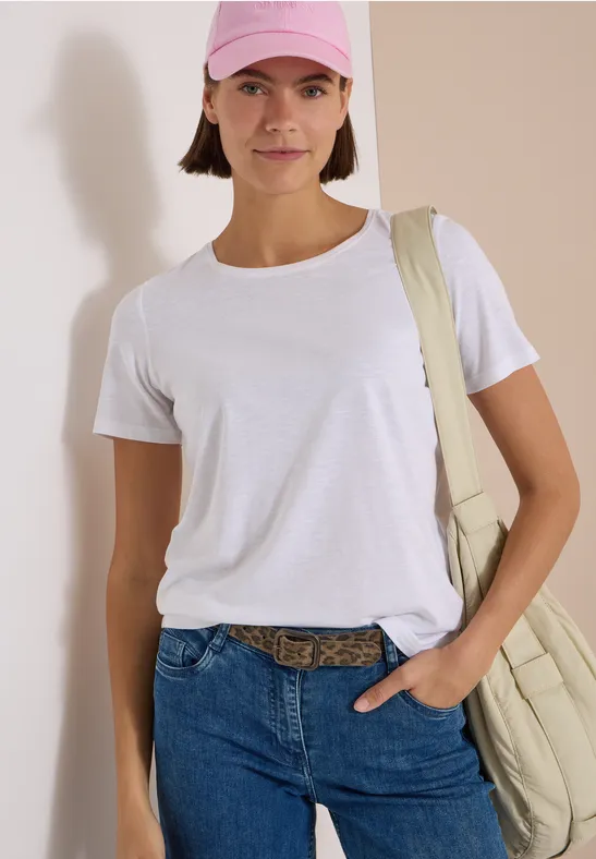 Frau in weißem T-Shirt, Jeans und rosa Cap mit Umhängetasche, stehend vor heller Wand.