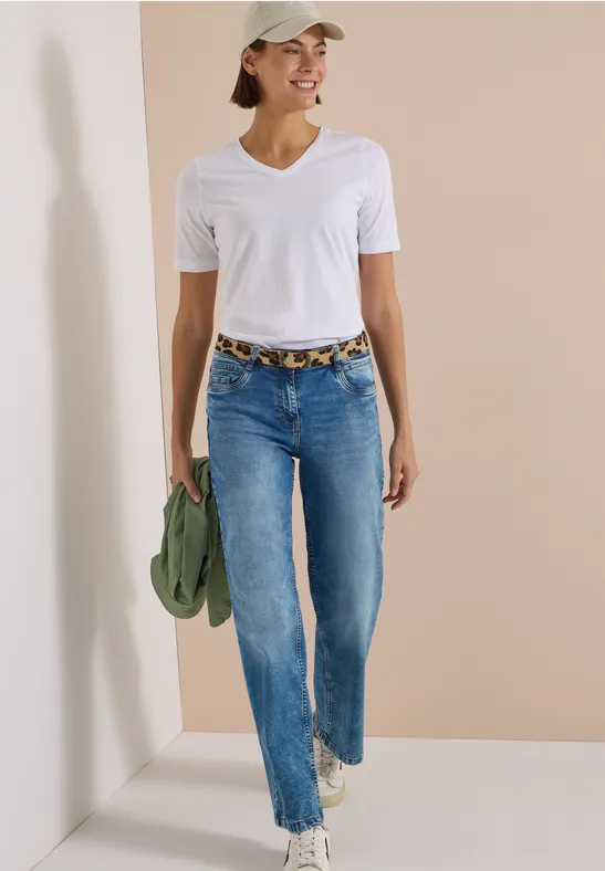 Frau in weißem T-Shirt, Jeans und Cap läuft lächelnd, hält grüne Jacke und trägt Leopardenmuster-Gürtel.