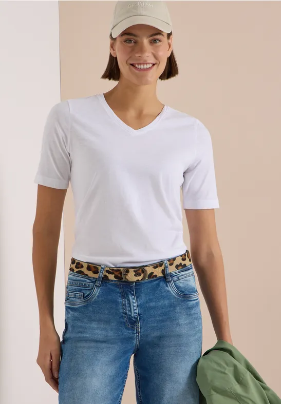 Frau in weißem V-Ausschnitt-T-Shirt und Jeans mit Leoparden-Gürtel, trägt Cap und hält grüne Jacke.