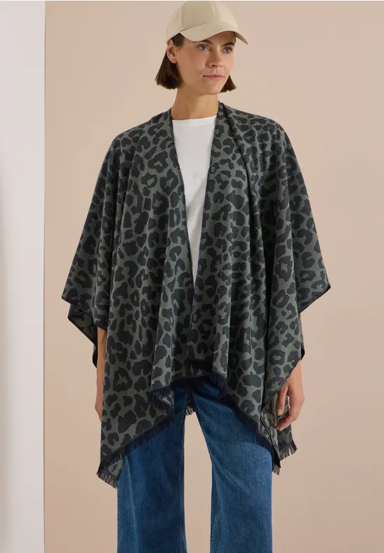 Frau mit gemustertem Poncho-Cardigan, weißem T-Shirt, Jeans und Baseballcap vor hellem Hintergrund.
