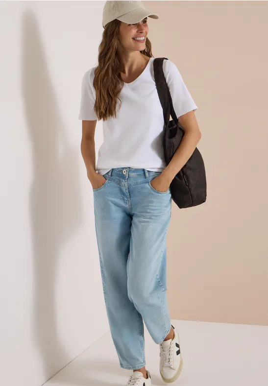 Vrouw in wit t-shirt, lichtblauwe spijkerbroek, sneakers en pet, schoudertas, neutrale achtergrond.