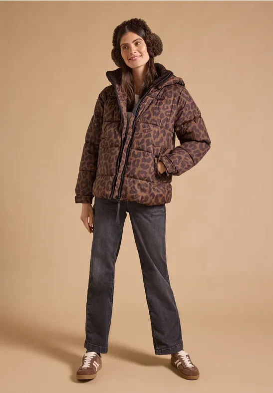Vrouw met krullen in bruine, gemêleerde gewatteerde jas, donkere jeans en witte schoenen voor een beige achtergrond.