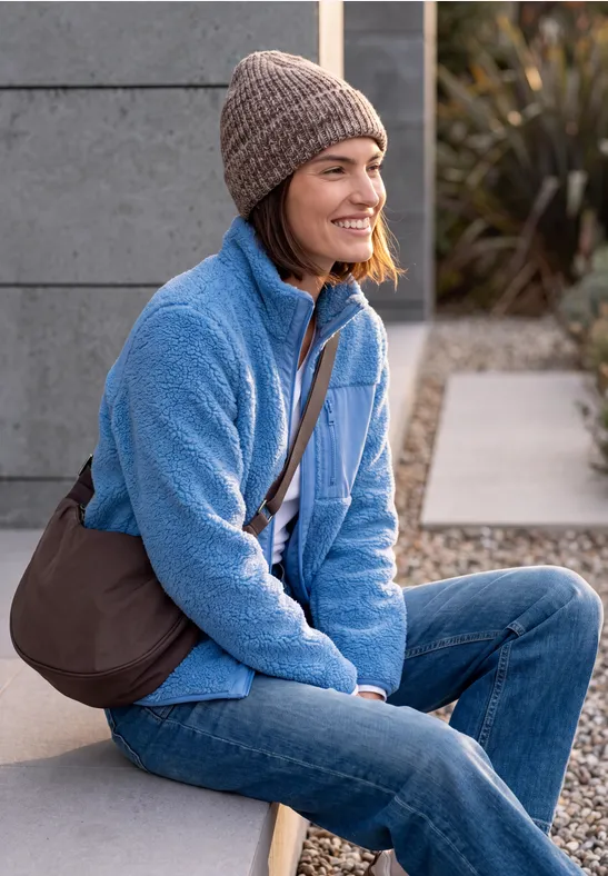 Frau sitzt draußen in blauer Fleecejacke, Jeans und Mütze mit Umhängetasche und lächelt.