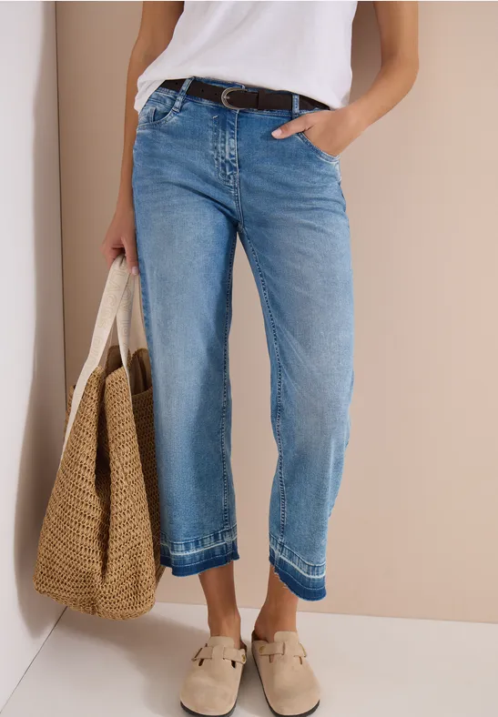 Person trägt weite blaue Jeans mit Gürtel, weiße Bluse, Korbtasche und hellfarbene Hausschuhe.