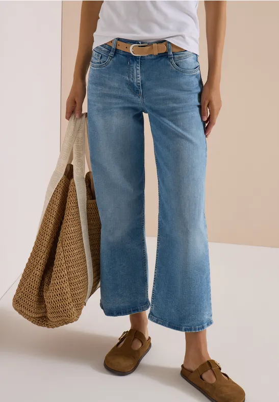 Person trägt weißes Shirt, weite hellblaue Jeans, braune Schuhe und hält eine Strohtasche.