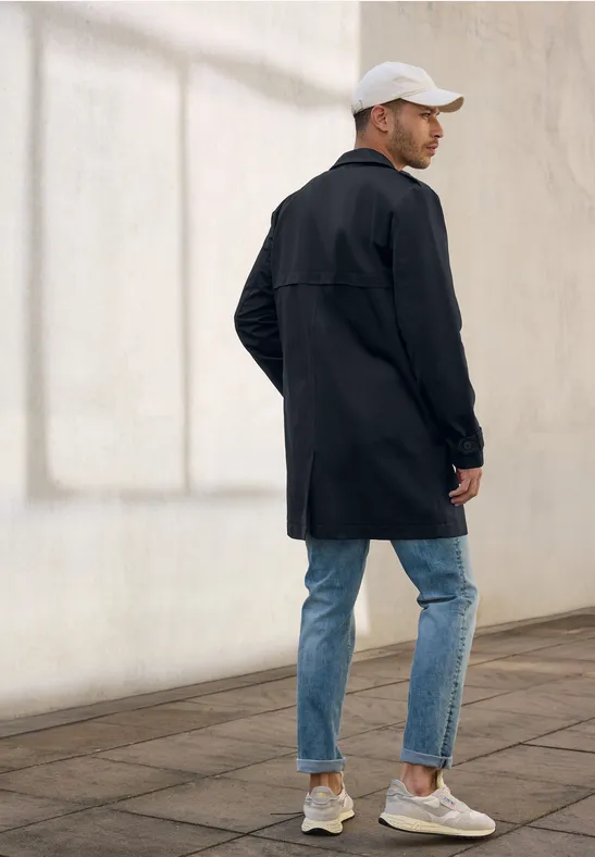 Man met zwarte jas, spijkerbroek en witte pet staat zijwaarts.