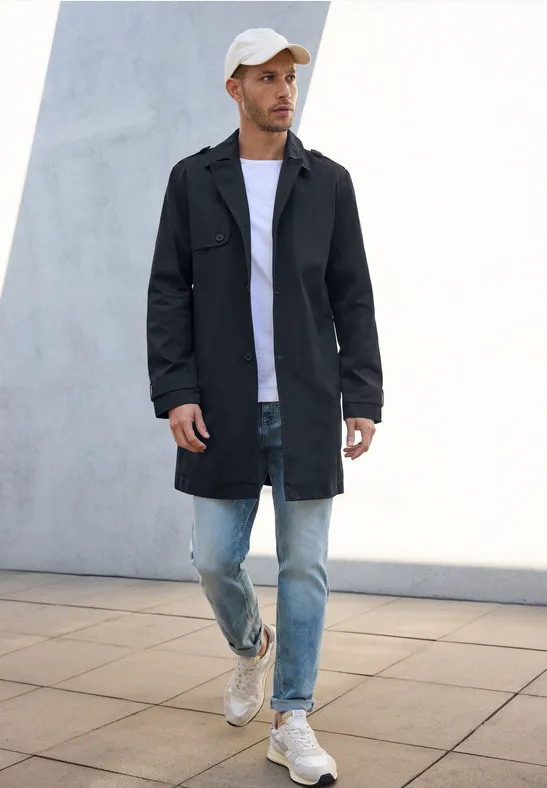 Man met zwarte jas, wit shirt, jeans en pet wandelt buiten.