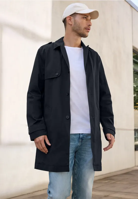 Man met zwarte jas, wit shirt, spijkerbroek en pet staat buiten.