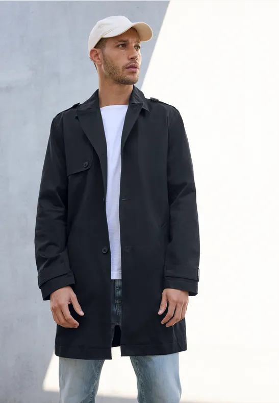 Man met zwarte jas, wit shirt en pet staat voor een lichte muur.