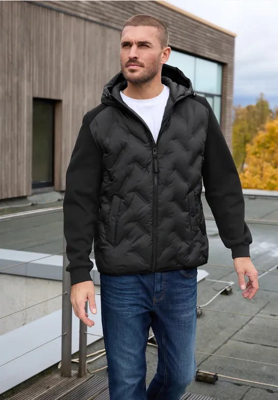 Un homme en veste matelassée noire, T-shirt blanc et jean bleu sur une terrasse avec façade en bois.