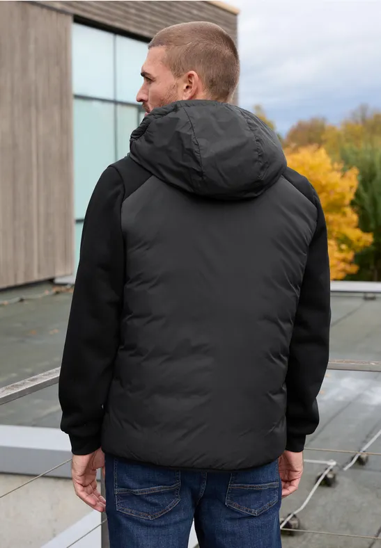 Homme vu de dos en doudoune noire et jean sur terrasse avec arbres d'automne en arrière-plan.