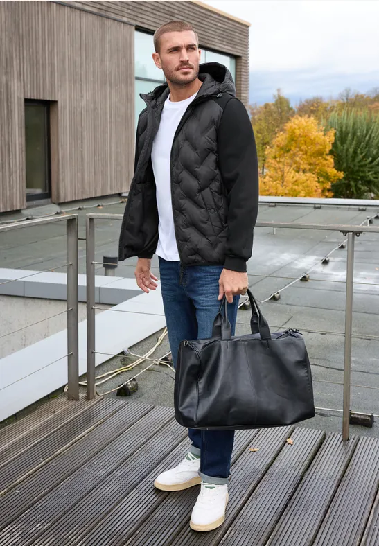 Un homme en doudoune noire, jeans et baskets blanches tient un sac de voyage noir sur une terrasse en bois.